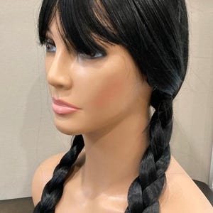 May include: A mannequin head with a black wig featuring blunt bangs and two long, braided pigtails. The wig is a solid black color and appears to be made of synthetic fibers. The mannequin has a neutral expression and is set against a neutral background.