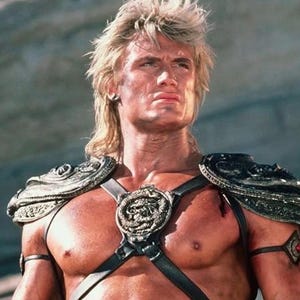 May include: A muscular person with blonde hair and a stern expression, wearing ornate silver and black armor. The armor includes shoulder pads, a chest piece with a central emblem, and armbands. The image has a vintage, action-movie aesthetic.