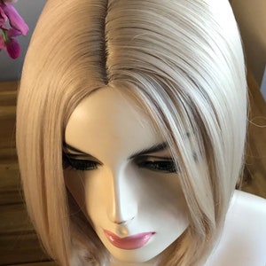 Peut inclure: Perruque blonde avec une raie au milieu, coiff&eacute;e en carr&eacute; mi-long. La perruque est pr&eacute;sent&eacute;e sur une t&ecirc;te de mannequin, mettant en valeur les cheveux lisses et droits et les reflets subtils. La couleur de la perruque est blond clair naturel.