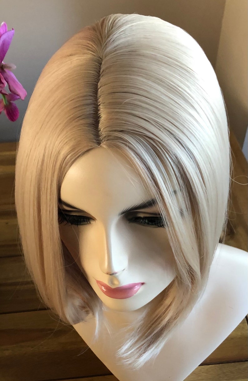 Peut inclure: Perruque blonde avec une raie au milieu, coiff&eacute;e en carr&eacute; mi-long. La perruque est pr&eacute;sent&eacute;e sur une t&ecirc;te de mannequin, mettant en valeur les cheveux lisses et droits et les reflets subtils. La couleur de la perruque est blond clair naturel.