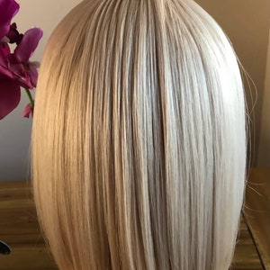 Peut inclure: Perruque blonde de style bob avec des cheveux raides et lisses. La perruque a un style naturel et est probablement faite de fibres synth&eacute;tiques. Les cheveux sont d'un blond clair et neutre avec des reflets subtils, et la coupe est un bob classique, au menton.