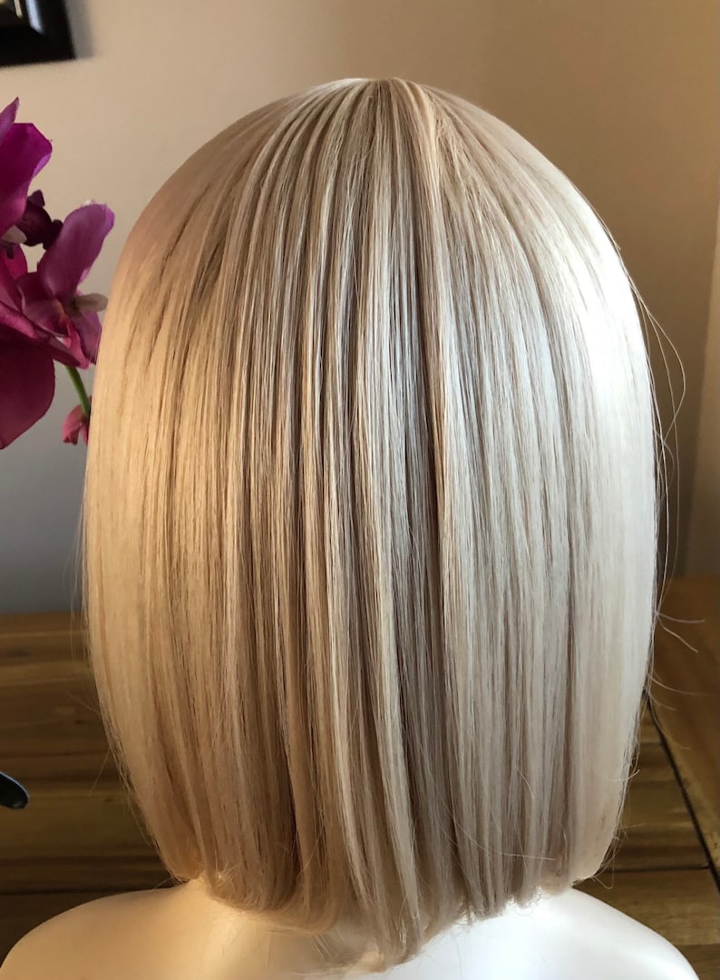 Peut inclure: Perruque blonde de style bob avec des cheveux raides et lisses. La perruque a un style naturel et est probablement faite de fibres synth&eacute;tiques. Les cheveux sont d'un blond clair et neutre avec des reflets subtils, et la coupe est un bob classique, au menton.