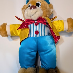 May include: A plush teddy bear dressed as a clown, wearing a blue satin hat, a yellow jacket with red and white polka-dot lapels, a red bow tie, and blue satin pants. The bear has a white and blue striped shirt with two red buttons.
