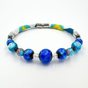Op de afbeelding: Een armband met een grote blauwe glaskraal in het midden, geflankeerd door kleinere blauwe, turquoise en doorzichtige glaskralen, afgewisseld met kleine zwarte kraaltjes. De band bevat een stoffen gedeelte met een geel, turquoise en wit patroon.