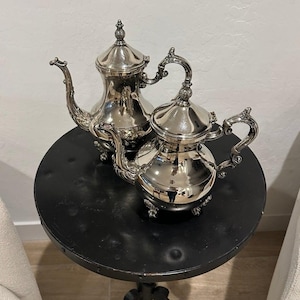 May include: Two polished silver-plated teapots with ornate, curved handles and spouts. Each teapot features a removable lid with a decorative finial and rests on four small, detailed feet. The metal surfaces are highly reflective and smooth.