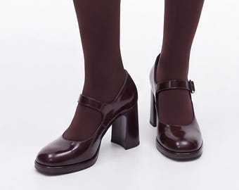 Op de afbeelding: Een paar donkerbordeauxrode Mary Jane-schoenen met een glanzende afwerking, voorzien van een ronde neus, een dikke blokhak en een enkel bandje over de wreef met gespsluiting, gedragen met ondoorzichtige donkerbordeauxrode panty's.
