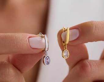 May include: Two drop earrings with oval-shaped stones. One earring is silver-toned with a light purple stone, and the other is gold-toned with a clear white stone. Both feature a hinged hoop closure and a small dangling oval charm.