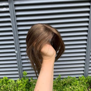 Puede incluir: Una peluca de color castaño medio con cabello liso y en capas, peinada con raya lateral. El cabello presenta reflejos sutiles y una textura natural, cayendo alrededor de la coronilla con mechones suaves que enmarcan el rostro.