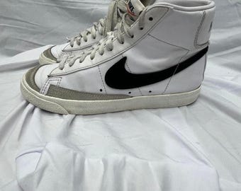 May include: A pair of white high-top sneakers featuring a prominent black Nike swoosh logo on the side, light gray suede overlays at the toe and heel, white laces, and a white rubber sole. The tongue displays a Nike brand tag with orange text.
