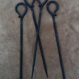 以下が含まれることがあります： ループ状のハンドルが付いた、手作りの暗い金属製の串が4本。串はハンドルの近くにねじれたデザインがあり、鋭い先端に先細りになっています。串は茶色の表面に配置されています。