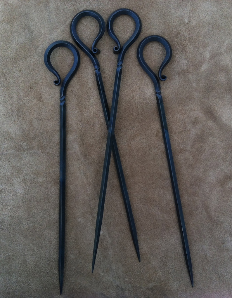 以下が含まれることがあります： ループ状のハンドルが付いた、手作りの暗い金属製の串が4本。串はハンドルの近くにねじれたデザインがあり、鋭い先端に先細りになっています。串は茶色の表面に配置されています。