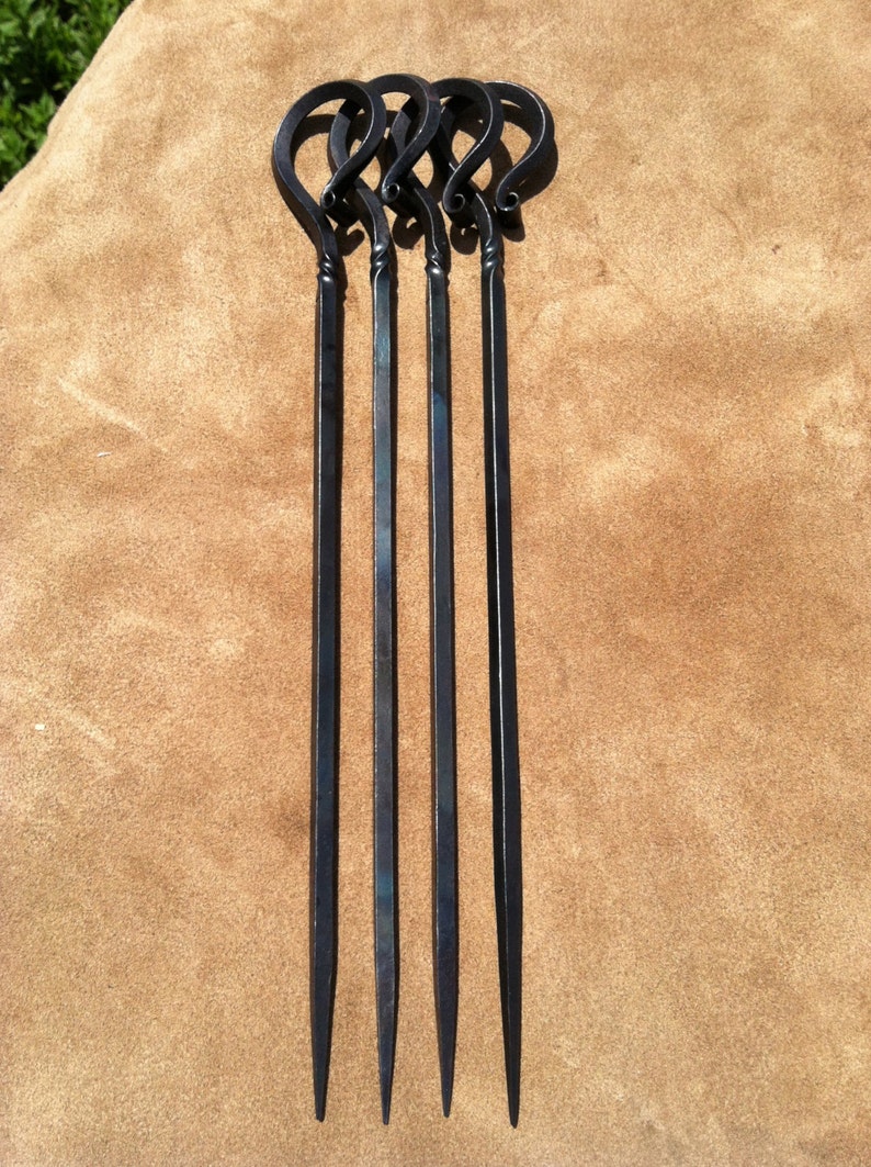 以下が含まれることがあります： 黒い金属製の串が4本。持ち手はループ状で、先端は尖っています。茶色の背景に垂直に配置され、洗練されたミニマルなデザインが特徴です。バーベキューに最適です。