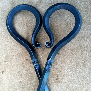 以下が含まれることがあります： 手作りの金属製の暖炉トング、ハート型のハンドル付き。トングは濃い青色で、ハンドルが交差する部分にねじれたデザインがあります。ハンドルの先端はカールしたデザインです。背景はタン色です。