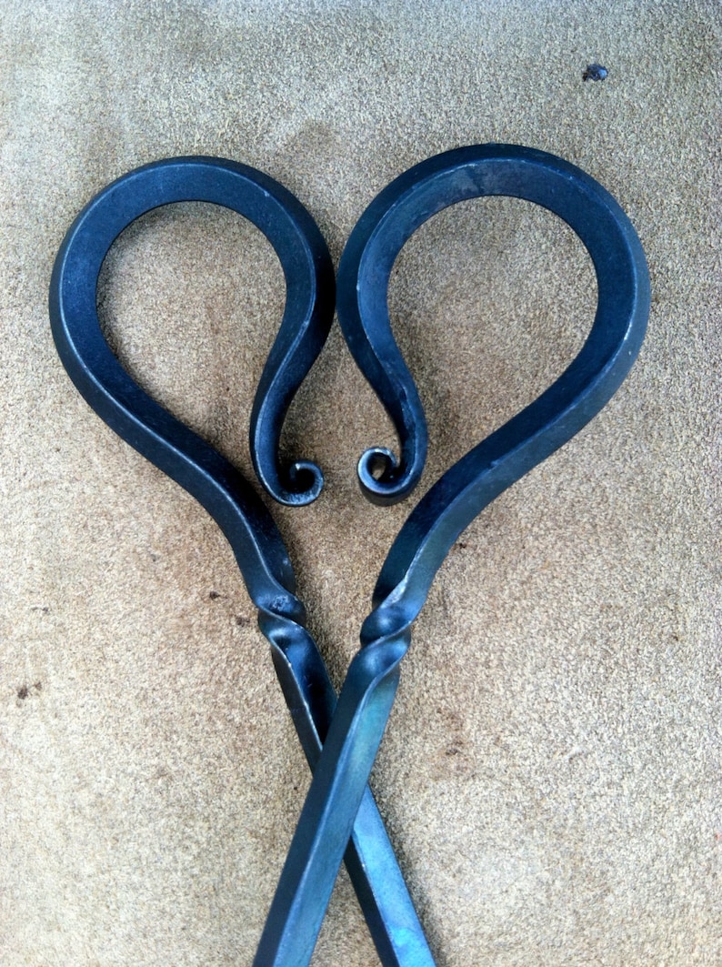 以下が含まれることがあります： 手作りの金属製の暖炉トング、ハート型のハンドル付き。トングは濃い青色で、ハンドルが交差する部分にねじれたデザインがあります。ハンドルの先端はカールしたデザインです。背景はタン色です。