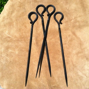 以下が含まれることがあります： 黒い金属製の串が4本。持ち手はループ状で、先端は尖っています。手作りのように見え、茶色の背景に配置されています。素朴な雰囲気やアウトドアを連想させます。