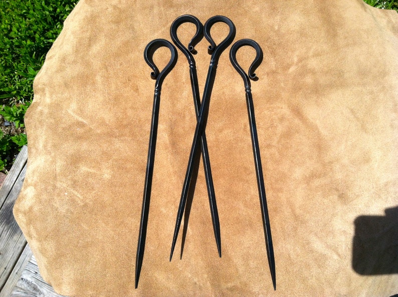 以下が含まれることがあります： 黒い金属製の串が4本。持ち手はループ状で、先端は尖っています。手作りのように見え、茶色の背景に配置されています。素朴な雰囲気やアウトドアを連想させます。