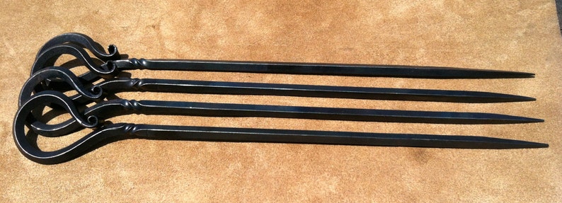 以下が含まれることがあります： 装飾的なループハンドルが付いた、黒い金属製の串が4本。串は長く細く、先端は尖っています。ハンドルは渦巻き模様の装飾的なデザインです。串は茶色の表面に配置されています。
