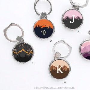 May include: Five phone ring holders with different mountain designs. Each ring holder has a different initial letter on it. The designs include a mountain range with stars, a mountain range with an orange sunset, a mountain range with a pink sunset, a mountain range with a purple sunset, and a mountain range with a brown sunset.