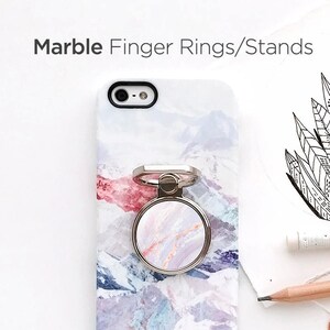 May include: A white phone case with a marble design and a silver phone ring holder. The ring holder is also marble patterned. The case is lying on a white surface with flowers and a pencil.