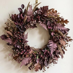 May include: A circular wreath constructed from a woven twig base, densely decorated with deep burgundy, plum, and brown dried foliage. The arrangement features assorted dried leaves, fern fronds, tall grasses, and clusters of small dark berries.