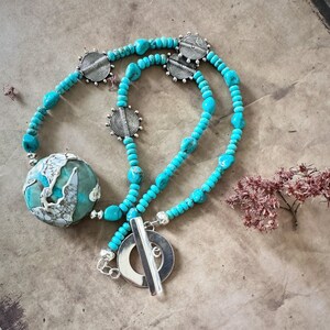 May include: A turquoise and silver necklace with a large turquoise pendant and a silver toggle clasp. The necklace is made of small turquoise beads and silver spacers.