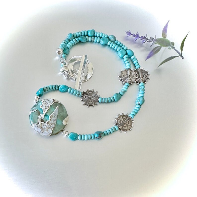 May include: A turquoise and silver necklace featuring a large turquoise pendant and silver accents. The necklace is made of small turquoise beads and silver spacers. The pendant is a large, round turquoise stone with a white pattern.