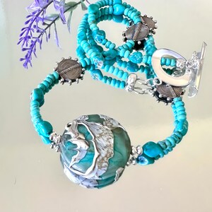 May include: A turquoise and silver beaded necklace with a large turquoise and silver pendant. The necklace is layered with multiple strands of turquoise beads and silver accents.