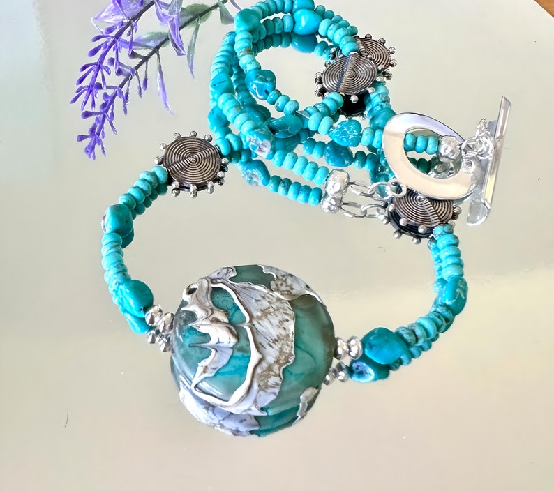 May include: A turquoise and silver beaded necklace with a large turquoise and silver pendant. The necklace is layered with multiple strands of turquoise beads and silver accents.