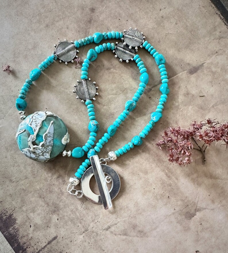 May include: A turquoise and silver necklace with a large turquoise pendant and a silver toggle clasp. The necklace is made of small turquoise beads and silver spacers.
