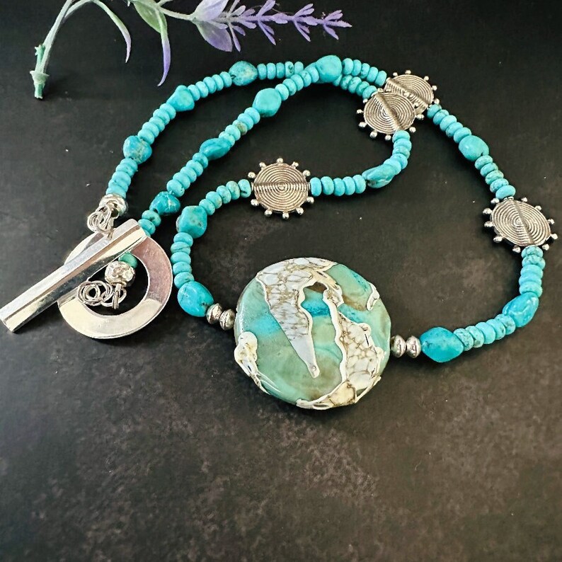 May include: A turquoise and silver necklace with a large, round turquoise pendant. The pendant has a white and grey marbled pattern. The necklace has a toggle clasp.