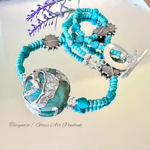 May include: A turquoise and silver necklace featuring a large turquoise and silver glass art pendant. The necklace is crafted with turquoise beads and silver spacers. The pendant is round and has a swirling design. The necklace is displayed against a white background.