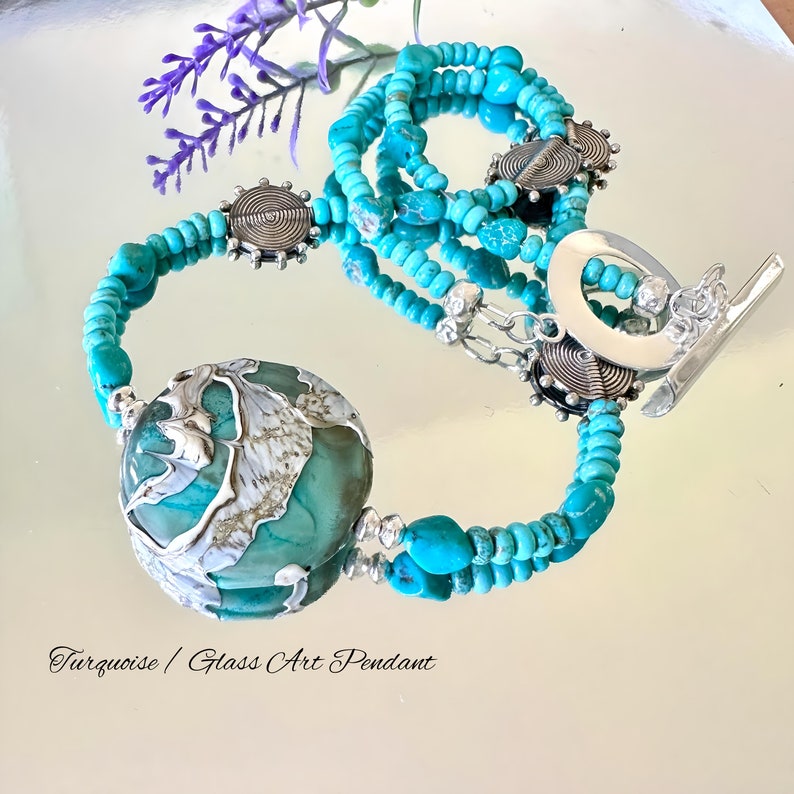 May include: A turquoise and silver necklace featuring a large turquoise and silver glass art pendant. The necklace is crafted with turquoise beads and silver spacers. The pendant is round and has a swirling design. The necklace is displayed against a white background.