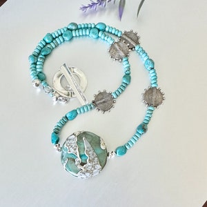 May include: A turquoise and silver necklace featuring a large turquoise pendant and silver circle charms. The necklace is made of small turquoise beads and silver beads.