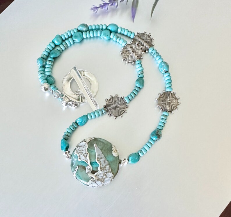 May include: A turquoise and silver necklace featuring a large turquoise pendant and silver circle charms. The necklace is made of small turquoise beads and silver beads.
