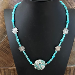May include: A turquoise and silver necklace with a large turquoise pendant. The necklace is made of small turquoise beads and silver spacers. The pendant is a round turquoise stone with a silver design.