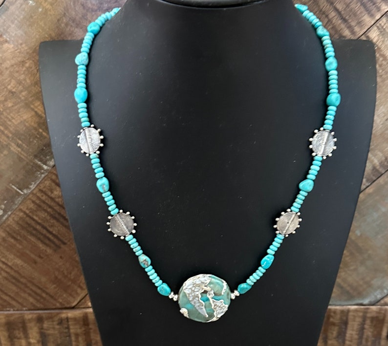 May include: A turquoise and silver necklace with a large turquoise pendant. The necklace is made of small turquoise beads and silver spacers. The pendant is a round turquoise stone with a silver design.