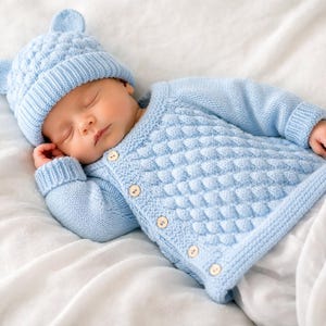 Puede incluir: Un bebé dormido con un conjunto de punto azul claro, que incluye un gorro con detalles de orejas y un cárdigan con botones. El bebé está acostado sobre una sábana blanca.