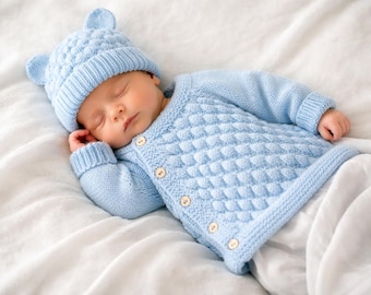 May include: A sleeping baby wearing a light blue knitted outfit, including a hat with small ear details and a button-up sweater. The baby is lying on a white sheet.