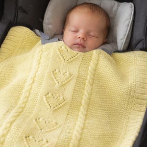 Op de afbeelding: Een slapende baby is gewikkeld in een zachte, gele gebreide deken met hartvormige uitsparingen en kabelbreiwerk. De baby zit in een zwart autostoeltje met een grijze hoofdsteun.