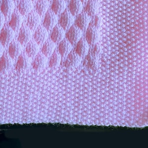 May include: A close-up of a soft, pink knitted blanket. The blanket features a textured diamond pattern in the center, surrounded by a smooth, ribbed border. The color is a light, pastel pink, perfect for a baby blanket or throw.