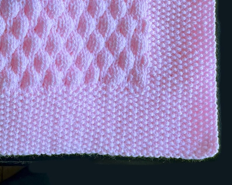 May include: A close-up of a soft, pink knitted blanket. The blanket features a textured diamond pattern in the center, surrounded by a smooth, ribbed border. The color is a light, pastel pink, perfect for a baby blanket or throw.