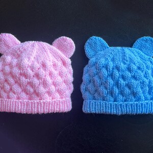 May include: Two knitted baby hats, one pink and one blue, each with a textured pattern and small ear details at the top. The hats are displayed on a dark surface.