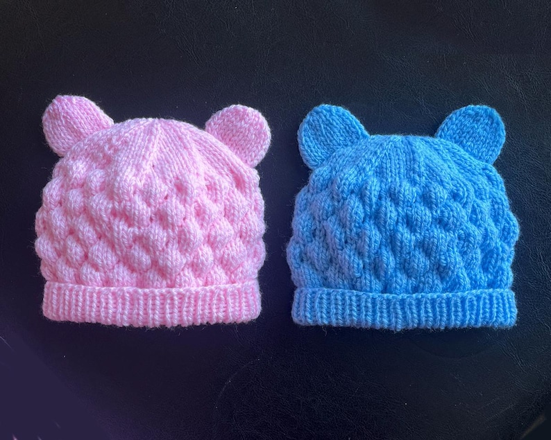May include: Two knitted baby hats, one pink and one blue, each with a textured pattern and small ear details at the top. The hats are displayed on a dark surface.