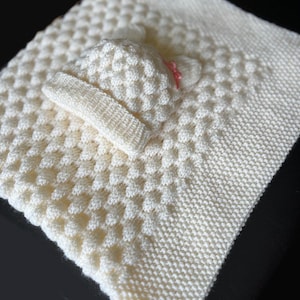 May include: A cream-colored, knitted baby blanket with a textured pattern. A matching hat and headband are placed on top. The hat has a small pink bow. The blanket is approximately 30 inches square.