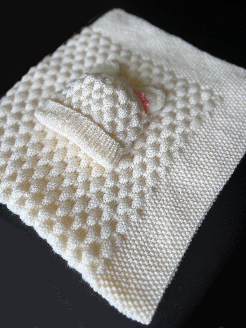 May include: A cream-colored, knitted baby blanket with a textured pattern. A matching hat and headband are placed on top. The hat has a small pink bow. The blanket is approximately 30 inches square.