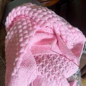 May include: A soft pink knitted blanket with a textured bubble stitch pattern. The blanket is folded, revealing different sections of the knitting, including a section with a lace-like design. A red knitting needle is visible.