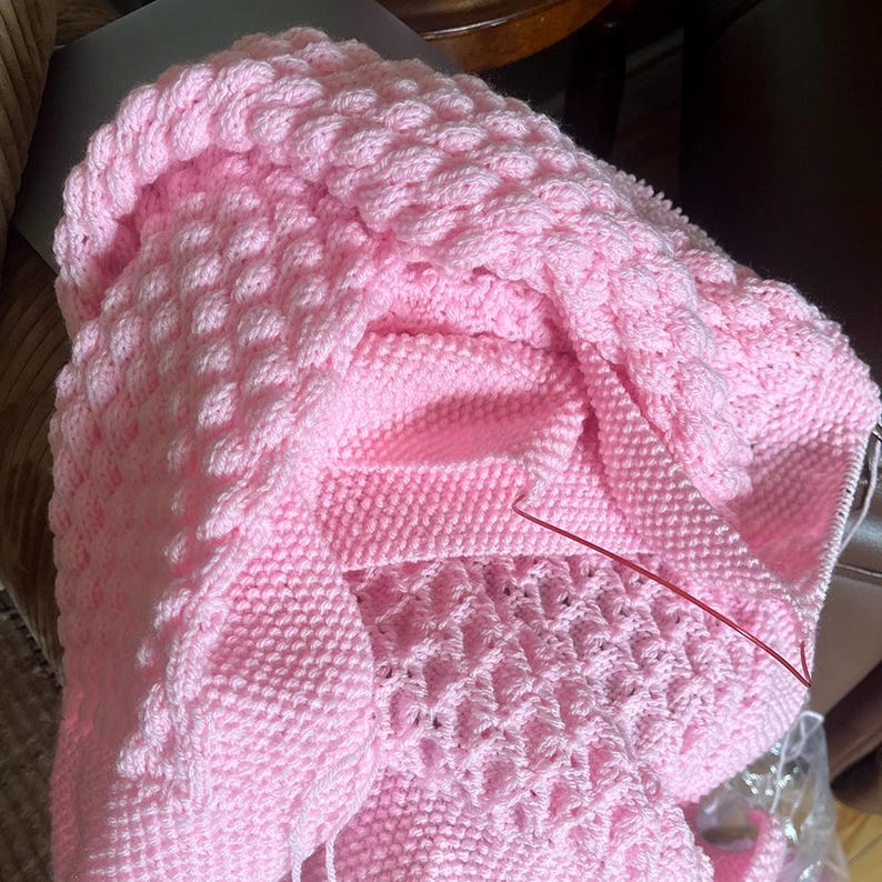 May include: A soft pink knitted blanket with a textured bubble stitch pattern. The blanket is folded, revealing different sections of the knitting, including a section with a lace-like design. A red knitting needle is visible.