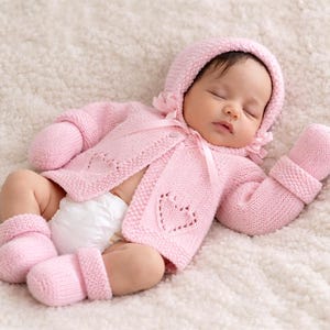May include: A sleeping baby wearing a pink knitted outfit, including a hooded jacket, mittens, and booties. The jacket features a heart design. The baby is lying on a soft, white, textured blanket.