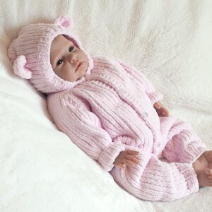 Puede incluir: Una mu&ntilde;eca beb&eacute; vestida con un mono de punto acanalado rosa claro y una capucha a juego con orejas de oso. La mu&ntilde;eca est&aacute; acostada sobre una manta blanca y esponjosa. El mono se abrocha con botones.