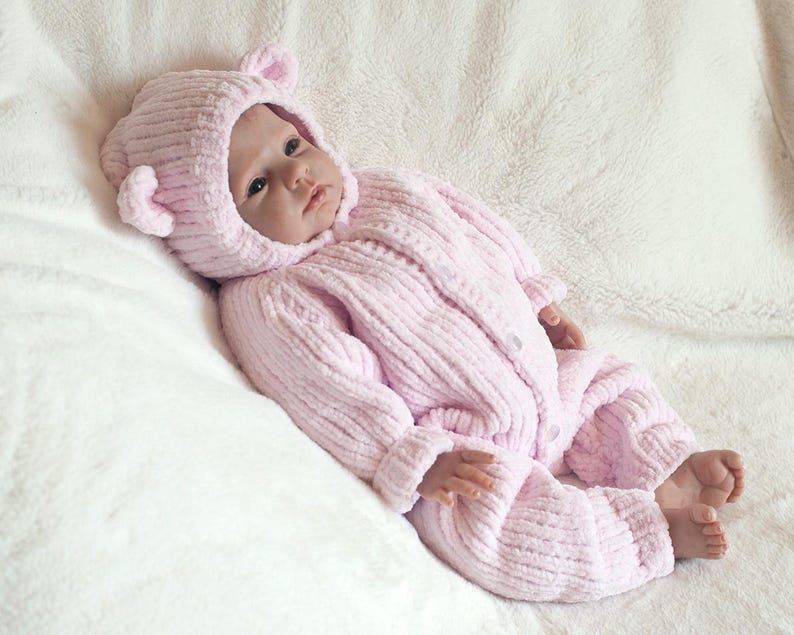 Puede incluir: Una mu&ntilde;eca beb&eacute; vestida con un mono de punto acanalado rosa claro y una capucha a juego con orejas de oso. La mu&ntilde;eca est&aacute; acostada sobre una manta blanca y esponjosa. El mono se abrocha con botones.